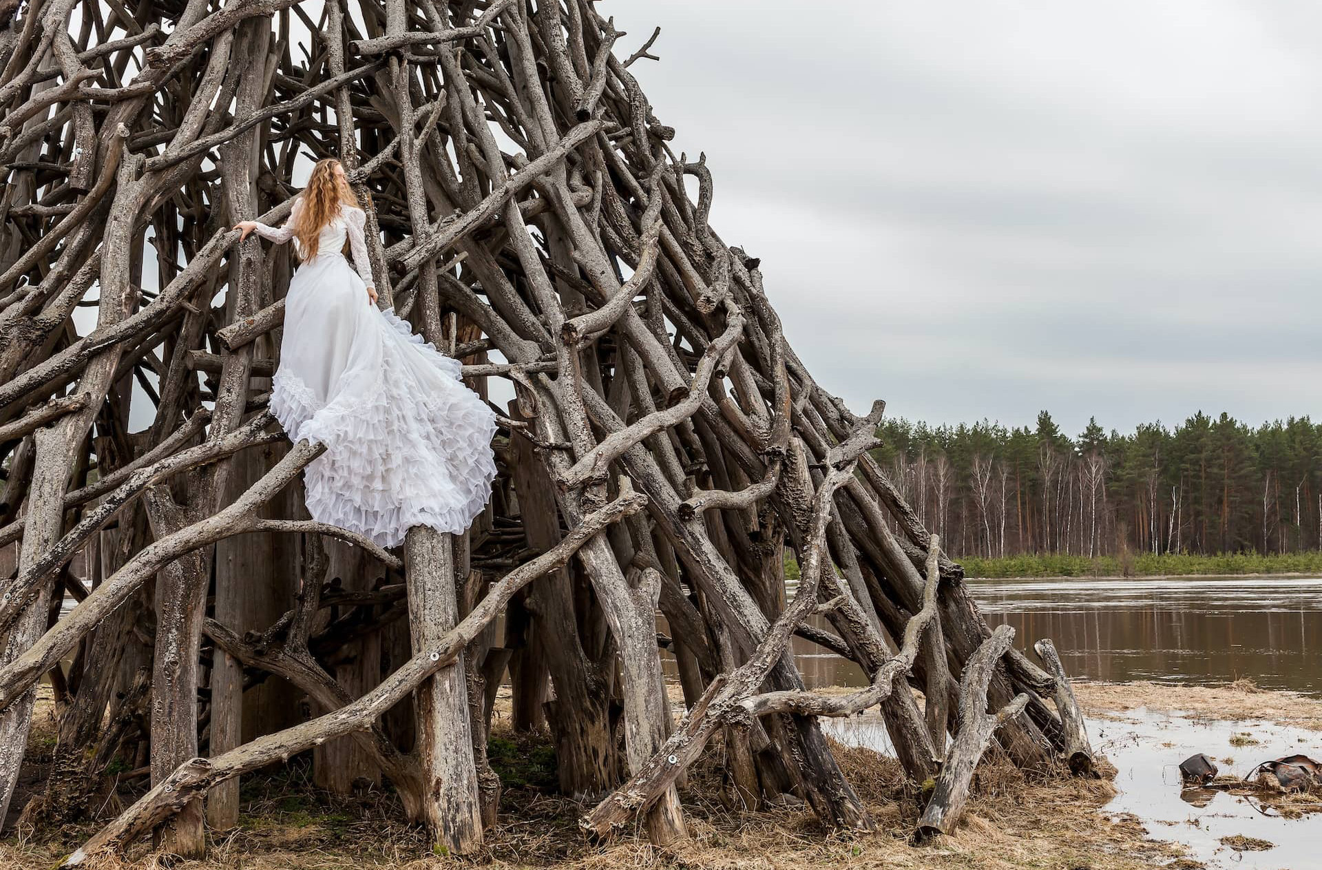 woman in wedding dress climbing up a tipi made out of driftwood next to a lake