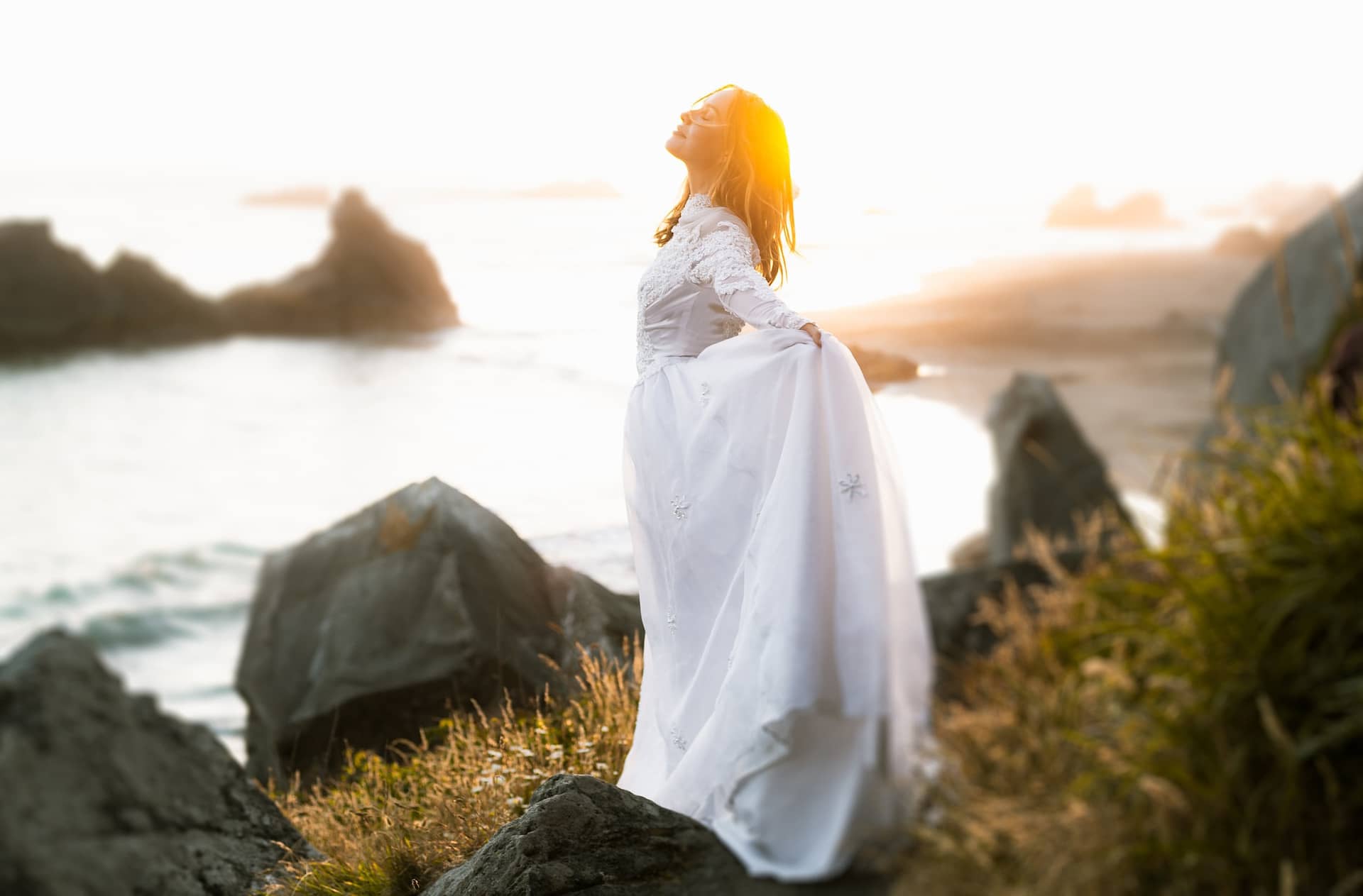 woman in wedding dress standing on a rocky cliff above the ocean as the sun sets