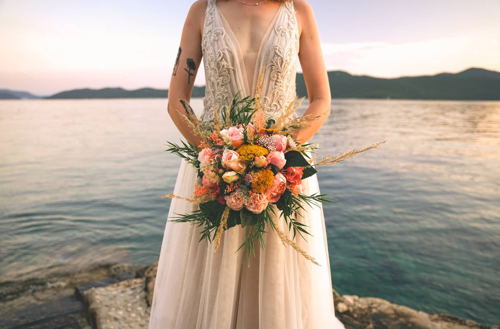 tatooed woman in wedding dress holding colorful boquet standing on rocks by a lake with mountains in the distance